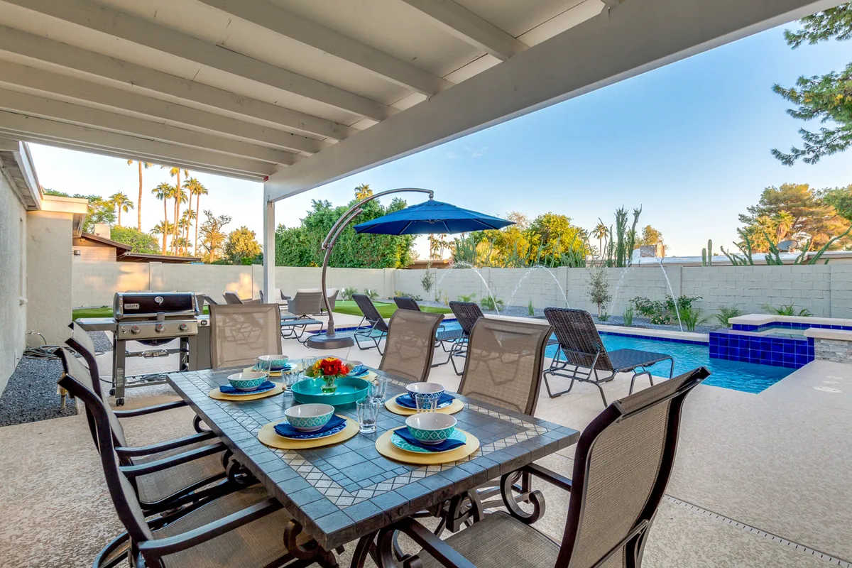 The image features an outdoor dining table set with blue plates, a sparkling swimming pool, an outdoor seating area, and a grill in the background. The scene is lively and cheerful, perfect for family meals and celebrations by the water.