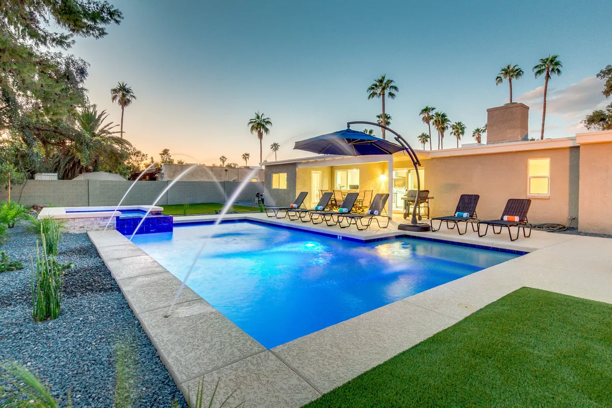 A tranquil backyard oasis featuring a sparkling swimming pool, poolside loungers, palm trees, and subtle evening lighting. The scene suggests an ideal setting for relaxing after a vibrant night out or enjoying a calm evening swim under the stars.