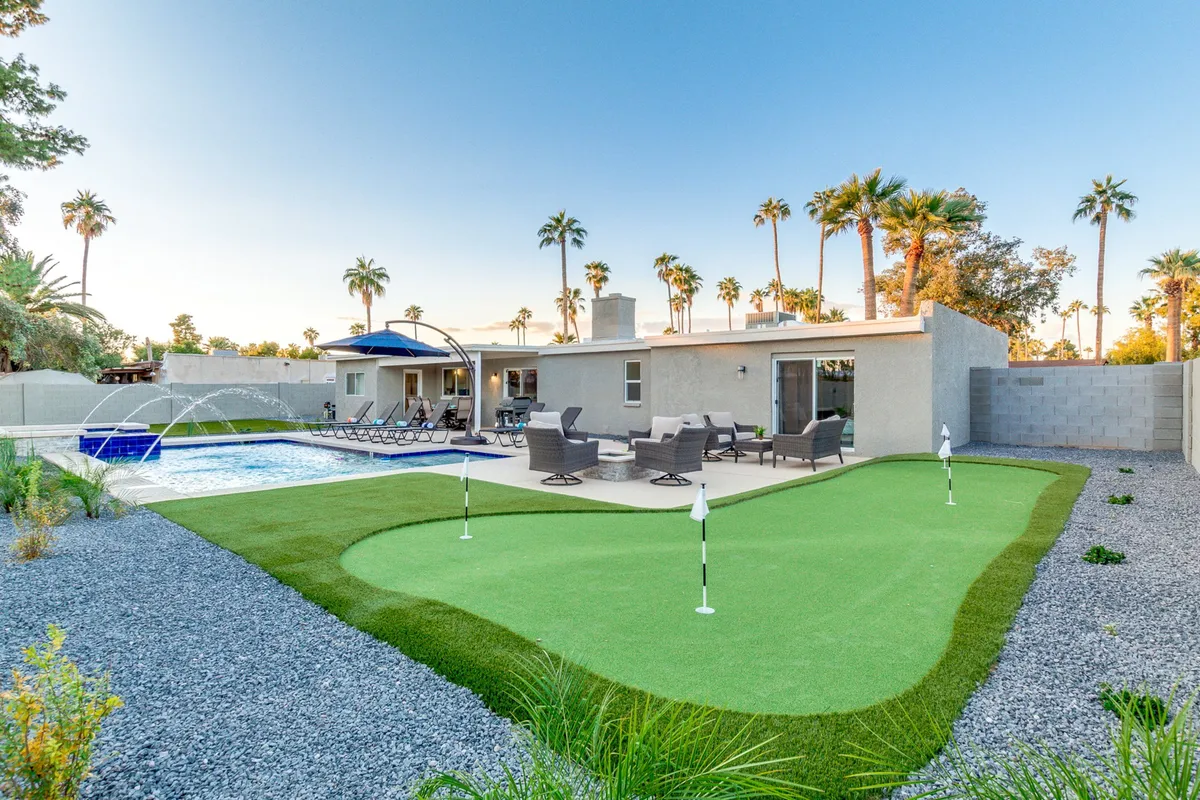 This exterior image prominently features a sparkling swimming pool, a lush putting green, several lounge chairs, and tall palm trees, all basking in sunlight. The layout encourages family-friendly activities like swimming and putting competitions, while the greenery and open sky evoke a sense of relaxation and fun.