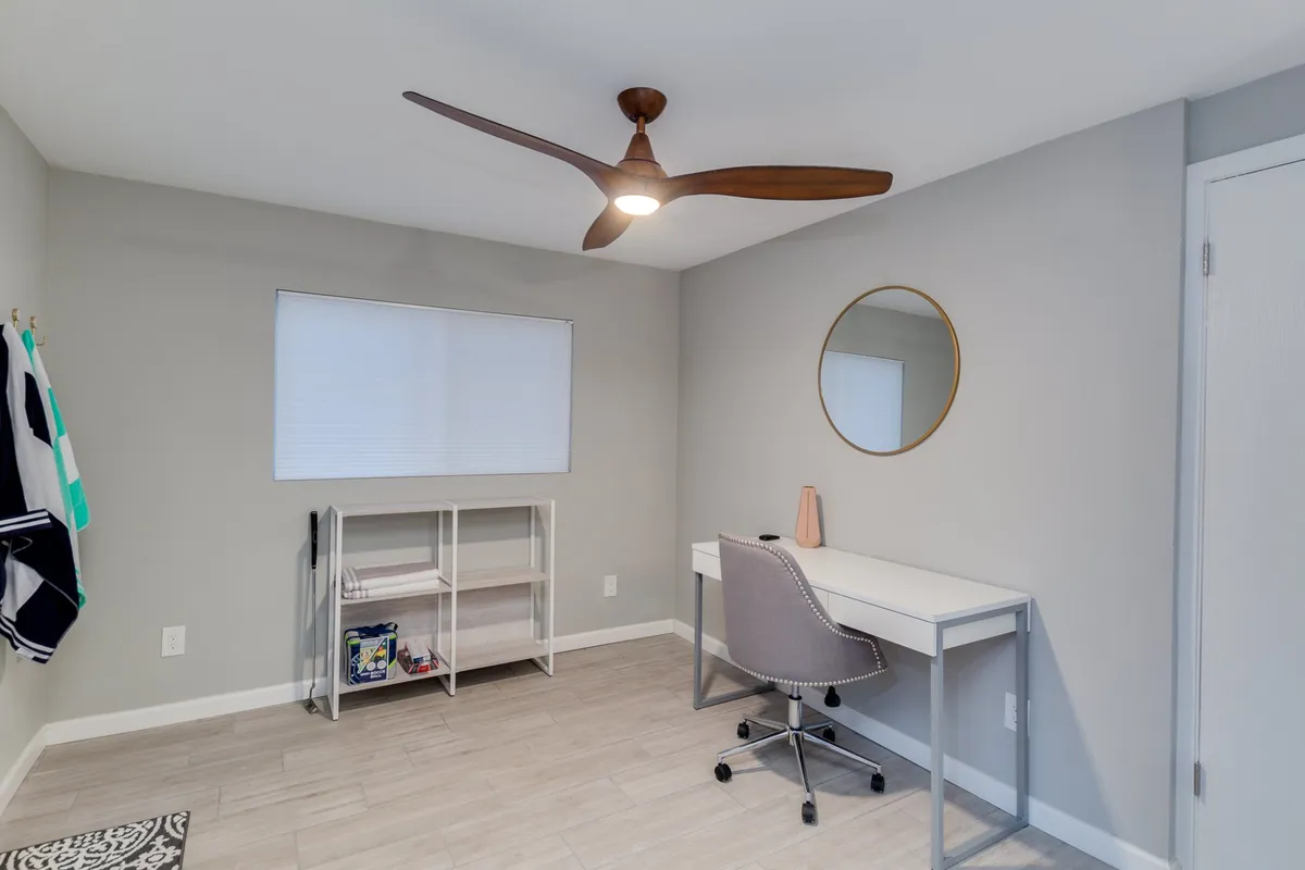 A bright, thoughtfully designed office space featuring a desk with a comfortable chair, a round mirror on the wall, a ceiling fan overhead, a window with blinds letting in natural light, and a practical shelving unit for organization. The uncluttered setup is ideal for focus and productivity.