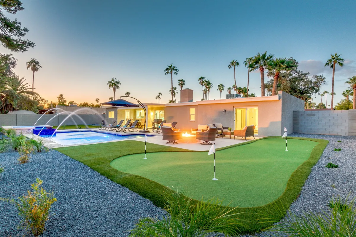 A wide-angle view of the backyard showcasing the sparkling swimming pool, a cozy fire pit area surrounded by seating, the lush putting green, and inviting lounge chairs framed by swaying palm trees. The setting is tranquil yet vibrant, with sunset lighting casting a warm glow over the entire outdoor escape.